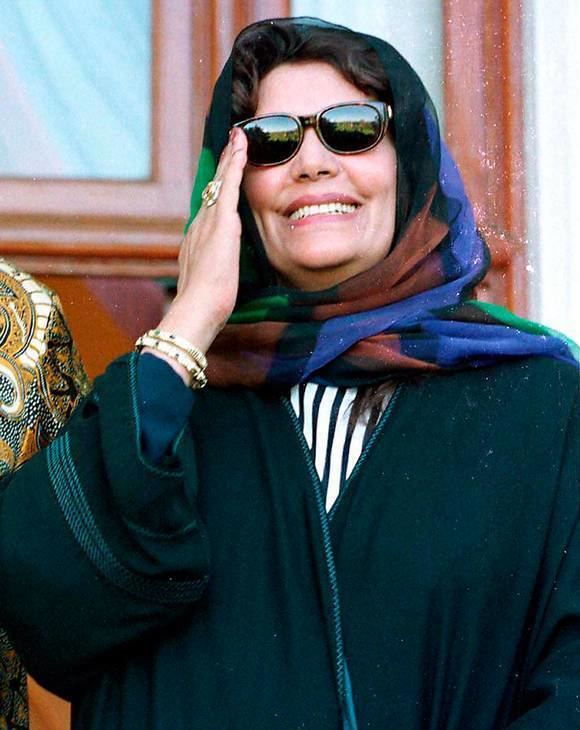 Safia Farkash smiling with her hand in her face and wearing sunglasses, a bottle green dress and a multicolored hijab.