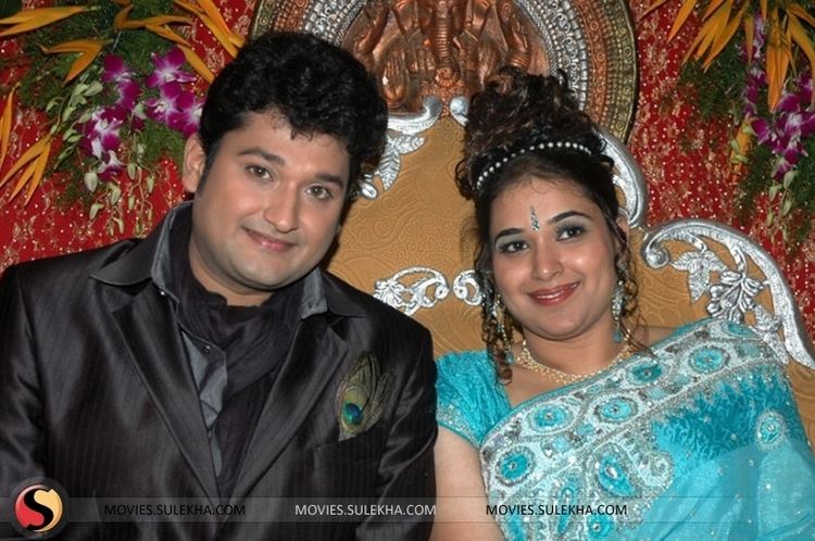 Sai Kiran smiling with curly hair and wearing a black coat with a peacock feather on its pocket and black scarf with his ex-wife Vyshnavi having a tika on the forehead, wearing a blue saree, necklace, a headband and dangling earrings