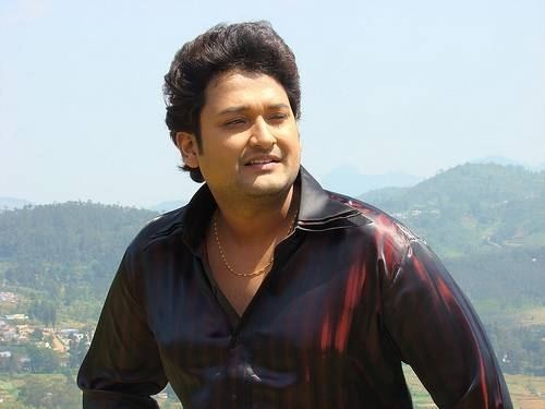 Sai Kiran smiling while looking afar with mountains in the background and wearing a maroon long sleeve glossy shirt and necklace