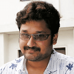 Sai Kiran smiling with a beard and mustache and wearing a white floral shirt and eyeglasses