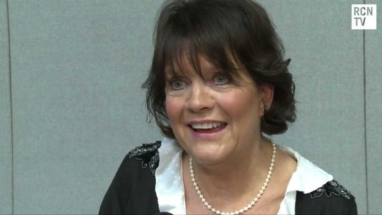 Sally Geeson smiling while wearing a black and white blouse and pearl necklace