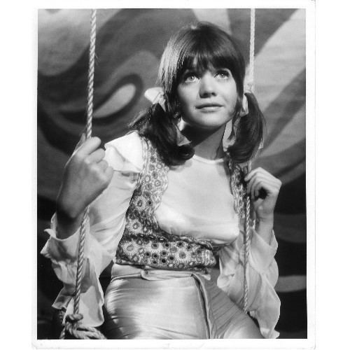 Sally Geeson looking upward while sitting on the swing and wearing white long sleeves and floral vest