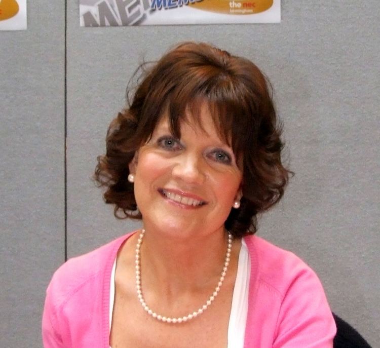 Sally Geeson smiling while wearing a pink blazer, pearl necklace and earrings