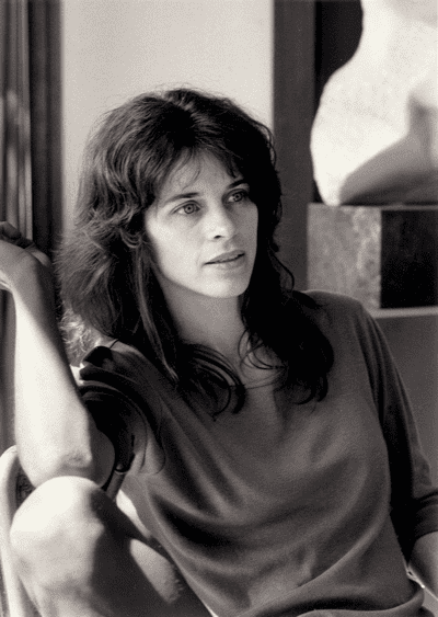 Sally Mann with black hair sitting down inside a house and wearing a gray shirt.
