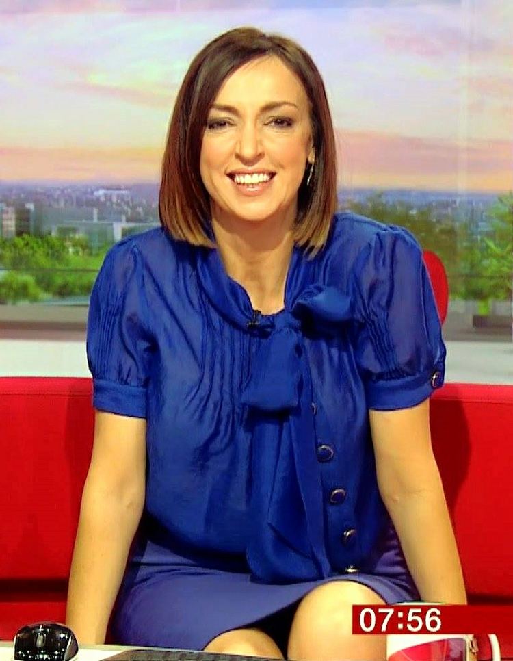 Sally Nugent smiling and sitting on a red couch at the BBC TV Studio while wearing a blue dress with a ribbon and earrings