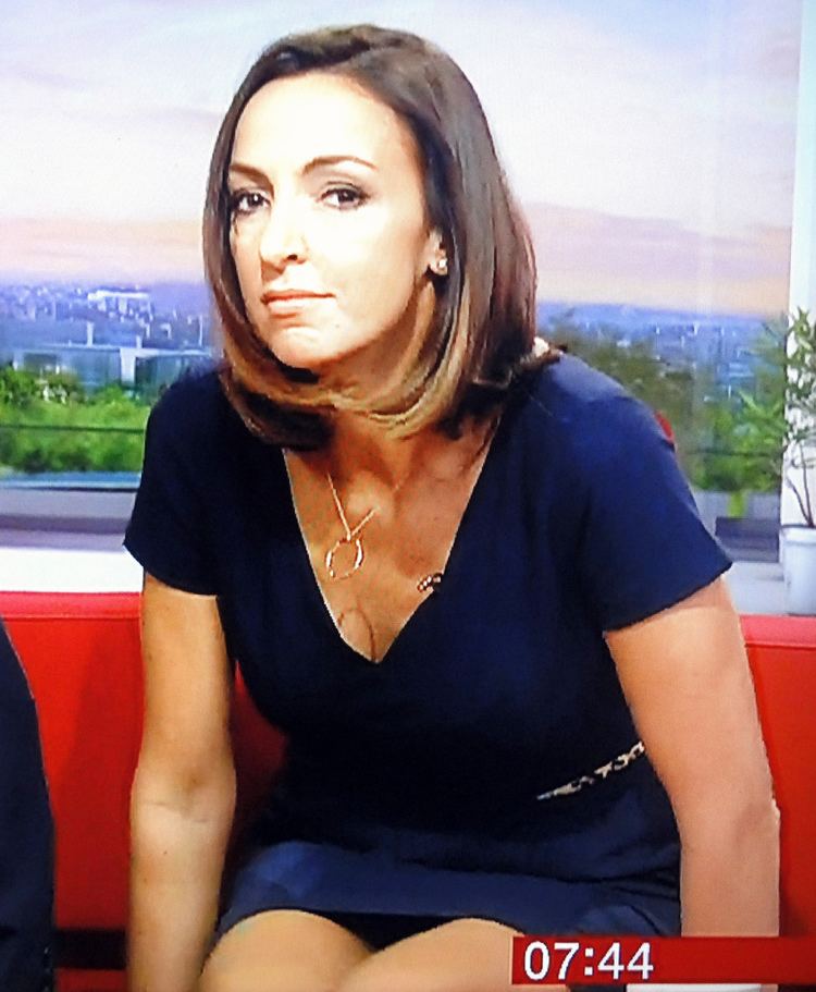 Sally Nugent sitting on a red couch at the BBC TV Studio while wearing a dark blue dress, necklace, and earrings