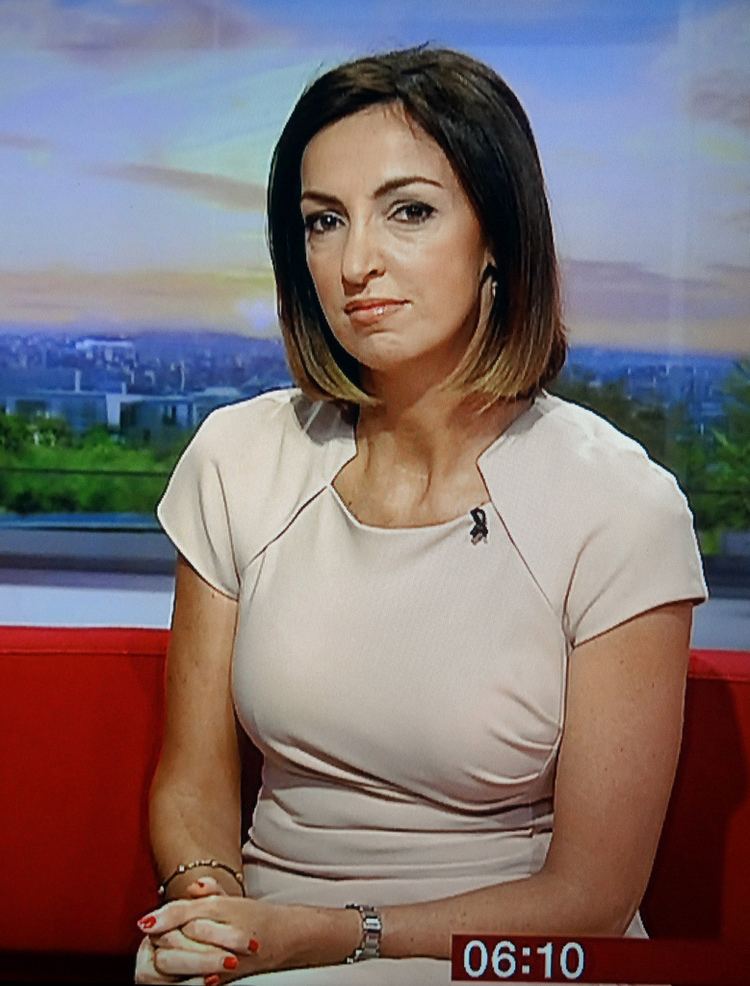 Sally Nugent sitting on a red couch at the BBC TV Studio while her hands together and wearing an off white dress, and bracelet
