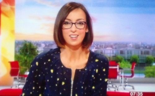 Sally Nugent reporting the news while wearing a blue and yellow blouse and eyeglasses