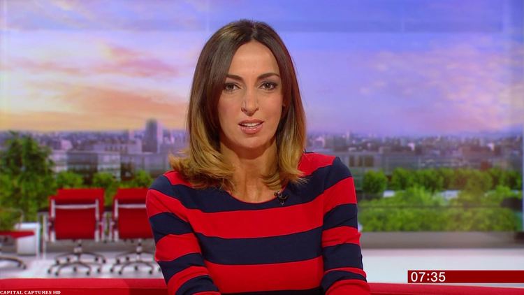 Sally Nugent reporting the news while wearing a red and black striped long sleeve blouse