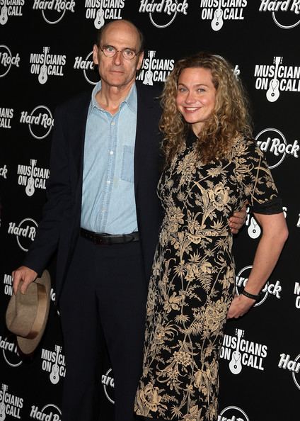 Sally Taylor with long curly hair and wearing a floral dress with her father, James Taylor wearing eyeglasses, a black suit, black pants, a light blue shirt, and holding a hat.