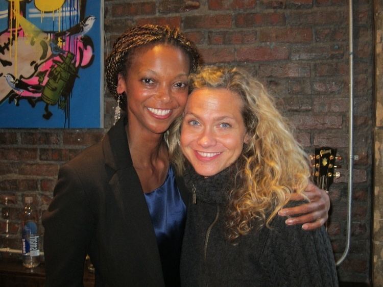 Sally Taylor smiling with curly blonde hair, wearing a black knitted sweatshirt with a lady wearing earrings, a black coat, and a blue shirt.