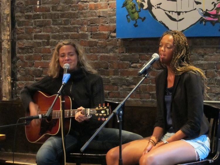 Sally Taylor with curly blonde hair, wearing a black knitted sweatshirt while playing guitar with a lady wearing a black coat, a blue shirt, and shorts while singing.
