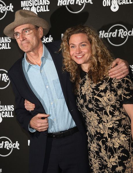 Sally Taylor with curly hair and wearing a floral dress while holding her father, James Taylor wearing eyeglasses, a brown hat, a black suit, black pants, and a light blue shirt.