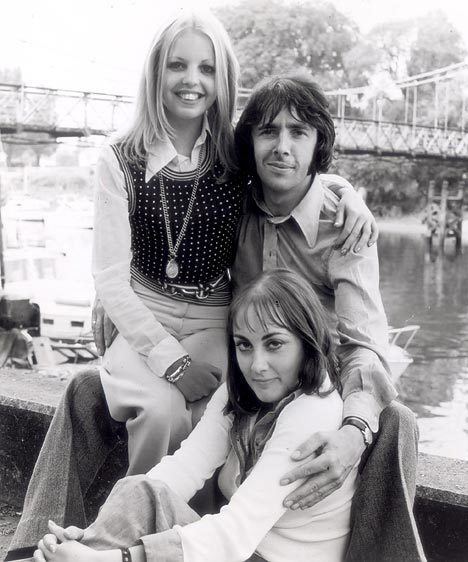Sally Thomsett smiling while sitting on the lap of Richard O'Sullivan holding Paula Wilcox on the shoulder with a serious face. Sally wearing a black vest and a white long sleeve while Richard is wearing a gray long sleeve, and Paula wearing a white long sleeve top.