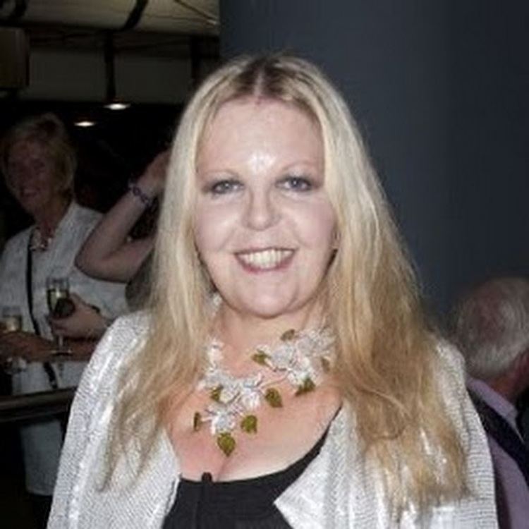 Sally Thomsett smiling while at her back is a woman looking at her wearing a white dress. Sally is with long blonde hair, wearing a necklace, a white blazer, and a black dress.