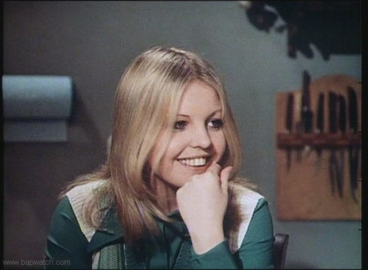 Young Sally Thomsett smiling while her hand on her chin, with blonde hair, and wearing a white and green long sleeve blouse.