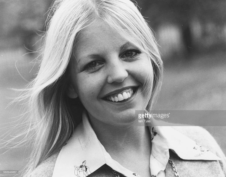 Sally Thomsett smiling, with blonde hair, wearing a necklace, and a knitted blouse with white collar.