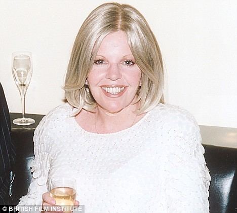 Sally Thomsett smiling while holding a glass of champagne, with blonde hair, wearing a necklace, and a white long sleeve blouse.