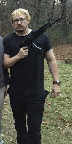 Sam Hyde standing and holding a rifle, with blonde hair, a mustache, and a beard, while wearing eyeglasses, a black t-shirt, black pants, and a wristwatch