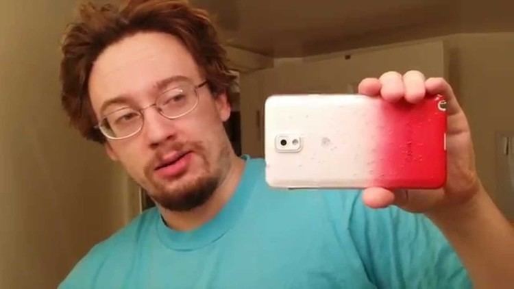 Sam Hyde holding and looking at his cellphone with a white and red case. Sam with a mustache and beard is wearing an aqua green t-shirt and eyeglasses