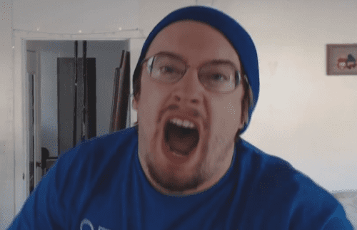 Sam Hyde shouting with an angry face, mustache, and beard while wearing a blue t-shirt, eyeglasses, and blue beanie