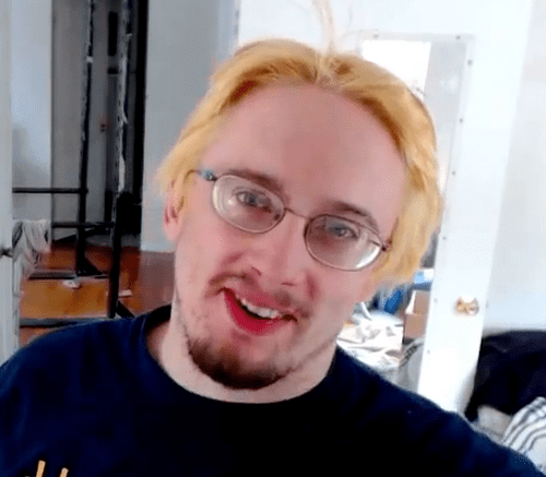 Sam Hyde smiling with blonde hair, mustache, and beard while wearing a black t-shirt and eyeglasses