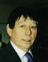Sammy Chung with a serious face, wearing a black coat over white striped long sleeves and a blue necktie.