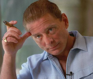 Sammy Gravano looking at something and scratching his forehead while holding a cigarette and wearing a sky blue polo shirt.
