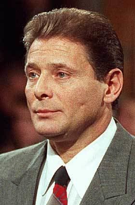 Sammy Gravano raising his hand for the sworn testimony at court wearing a gray suit as well as a black and red tie.