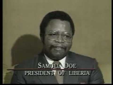 Samuel Doe giving a speech while wearing a black coat, white long sleeves, brown necktie, and eyeglasses