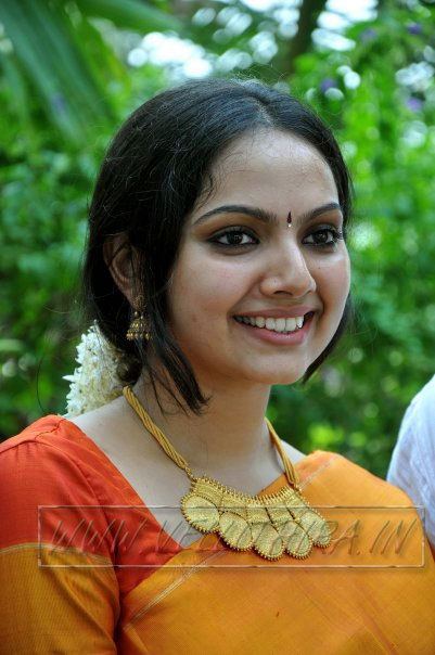 Samvrutha Sunil smiling, wearing earrings, necklace, and yellow-orange Indian traditional clothing.
