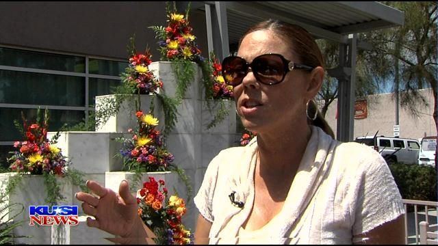In KUSI NES, Wendy Flanagan is serious, talking, hand in front open, behind is a white marble for funerals of the victims with flowers on top, she has long brown hair wearing a black sunglasses, gold hoop earrings, and a cream white top.