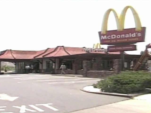 The San ysidro Mcdonald's way back in 1991, has red colored bricks and red roofs with Mcdonald’s name on top, in front is the parking with white lines and exit sign, at the right is Mcdonald’s large Road sign in and green bush in front.