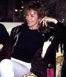 Sara Allen smiling while sitting on a couch and wearing black jacket and white pants