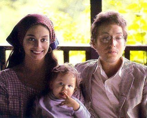 Sara Dylan in her white and pink dress while carrying their baby wearing violet sweatshirt and Bob Dylan in his stripe coat, white long sleeves and eyeglasses