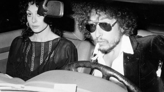Sara Dylan inside the car with Bob Dylan wearing shades, coat and long sleeves