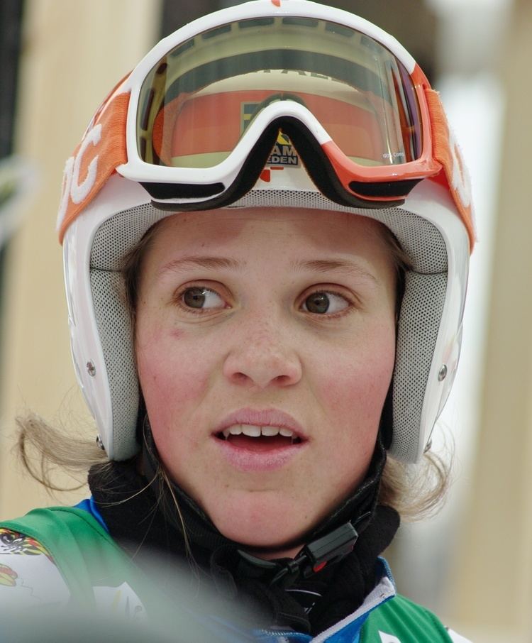 Sara Hector with a surprised facial expression, wearing a protective helmet and sunglasses, and a multi-colored jacket.