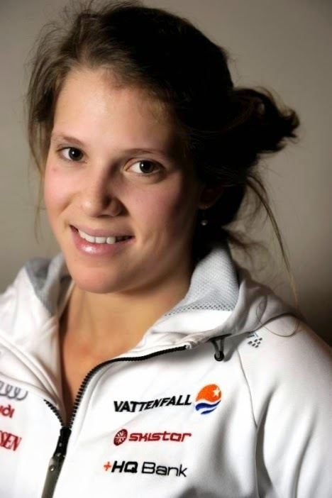 Sara Hector smiling, with messy hair, wearing pearl earrings and a white hoody jacket with logos.
