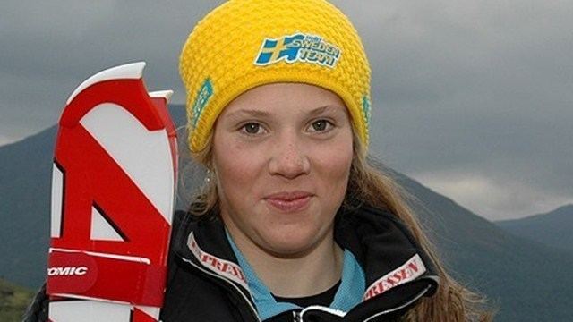 Sara Hector is smiling while holding a red and white ski, with long blonde hair, wearing earrings, a yellow bonnet, and a black jacket over a light blue and black shirt.