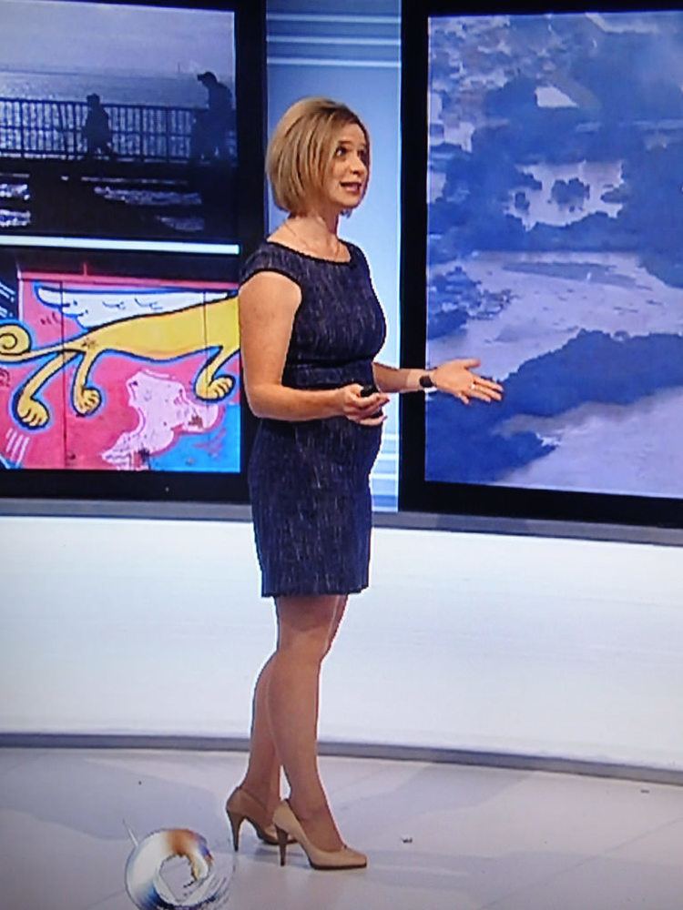 Sarah Keith-Lucas reporting the weather forecast while wearing a blue dress, wristwatch, and beige heels
