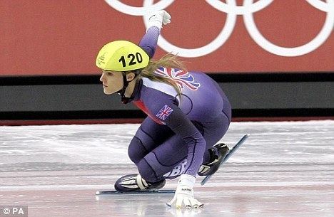 Sarah Lindsay skating while touching the ice rink, with a serious face, and wearing a yellow helmet with a number 120 print, a white and purple figure skates, white gloves, and UK Flag on her arm in a purple long sleeve full bodysuit