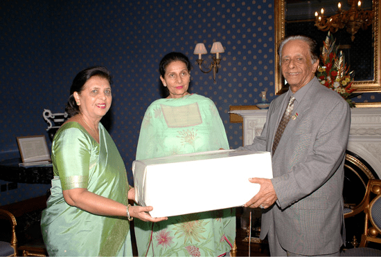Sarojini Jugnauth wearing a mint green dress and a bracelet while holding a gift for Sir Anerood Jugnauth wearing a gray suit and a tie with Preneet Kaur also wearing a mint green dress.