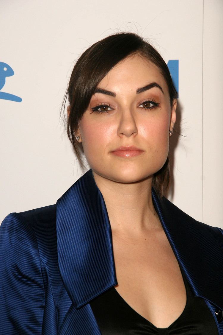 Sasha Grey smiling and wearing a black blouse under a blue coat.