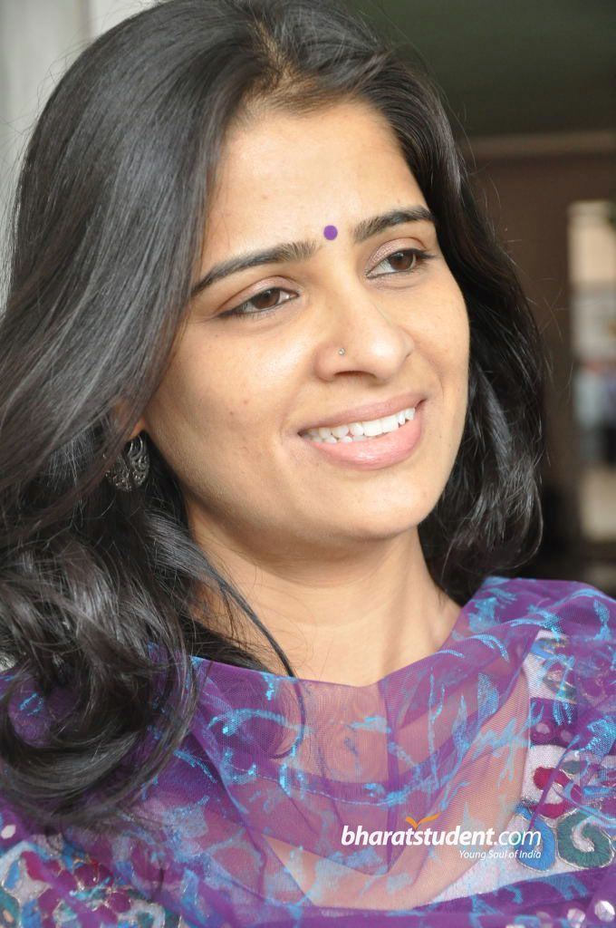 On the left, Satya Krishnan is smiling, standing, looking at her left, has black wavy hair, a bindi, and nose piercing, wearing earrings, a white printed shirt under a purple see through scarf, at the bottom right is a word written “bharatstudent.com” “Young Soul of India”