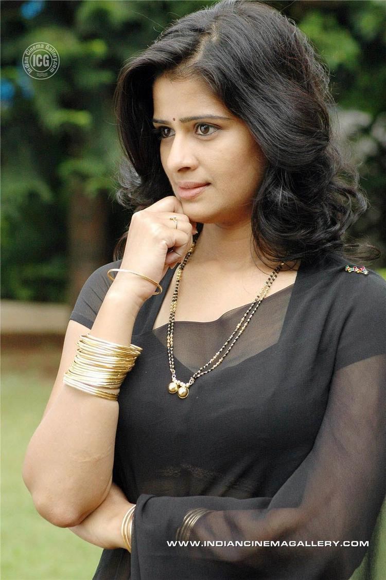 Satya Krishnan is serious, standing with his right hand closed under her chin, has long wavy hair, and a bindi on her forehead, wearing a gold bracelet and gold necklace with gold pendant, and a black see through saree.