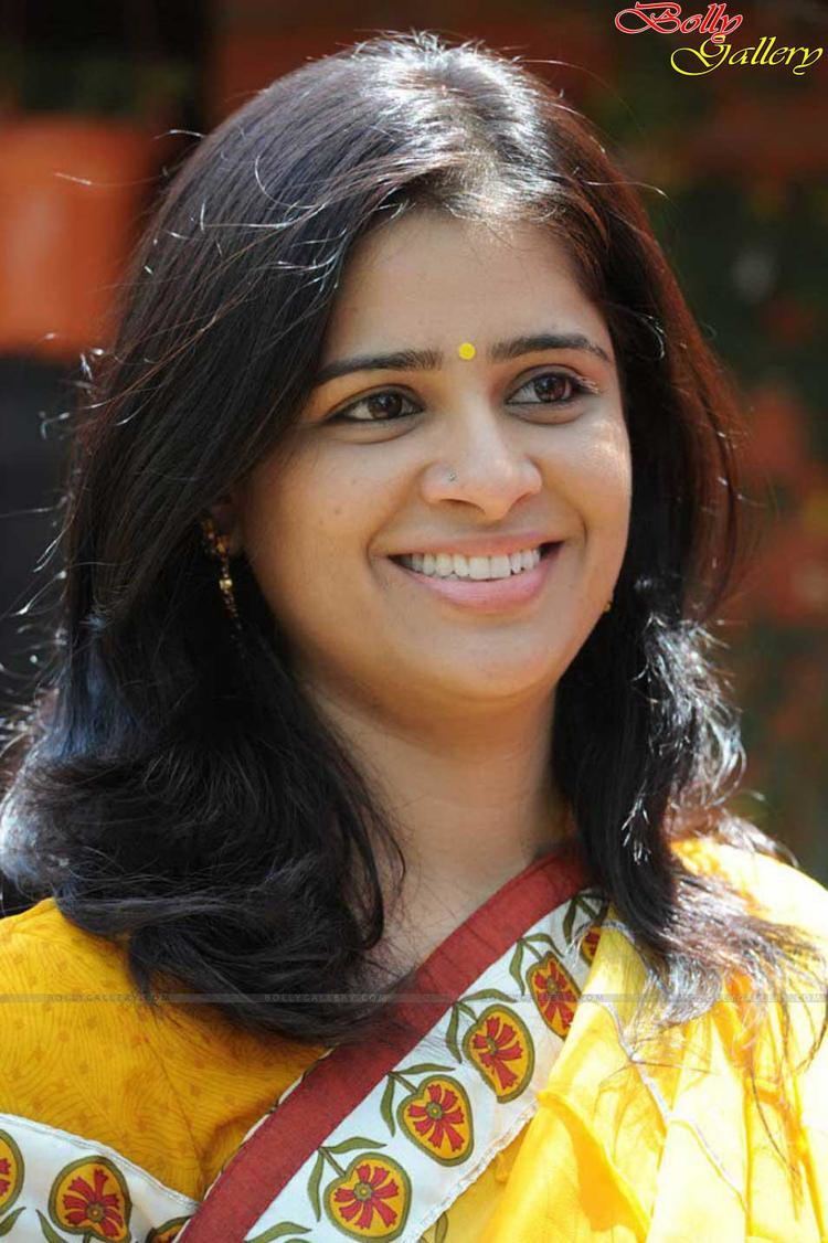On the left, Satya Krishnan is smiling, standing, looking at her left, has long black hair, a yellow bindi, wearing earrings, nose piercing and a yellow flower printed saree, At the top right is a word written “BollyGallery”