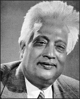 Satyendra Nath Bose is smiling has white hair, wearing white long sleeves, a black necktie under a gray suit.
