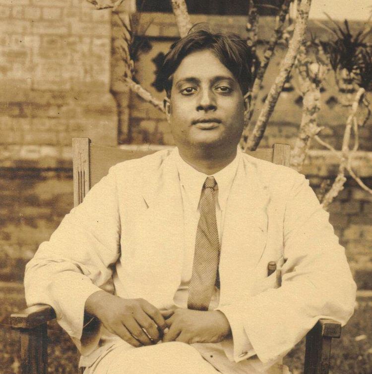 Satyendra Nath Bose is serious, has black hair, a mole on his right lower cheek, sits down on a brown chair, both hands holding each other, and a pen inside his pocket, wears white long sleeves, a brown necktie under a white suit with pocket, and white pants.