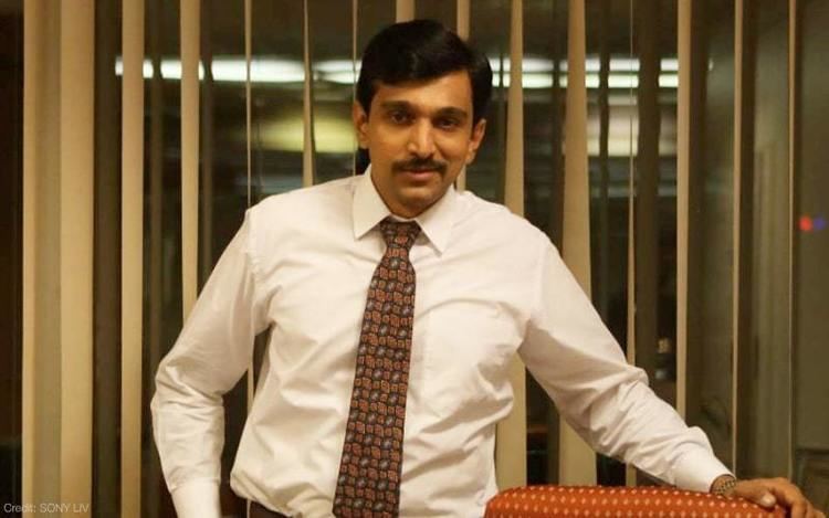 Pratik Gandi as Harshad Mehta smiling with a mustache and holding into a couch and wearing a white sleeve with a brown necktie