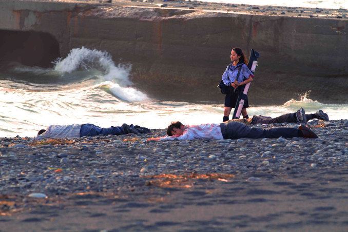 In the movie scene SchoolGirl Apocalypse 2011, Higarino is walking at a bay with three dead bodies a man from the left wearing long striped polo, jeans and shoes, in the middle a man wearing white long sleeve with blood, pants and a pair of shoes, a man on the right wearing pants and a pair of shoes. while holding her bag by her shoulder and using both hands to hold her sword covered with cloth, has long black hair, wearing white seifuku school uniform, black knee level socks and black skirt.